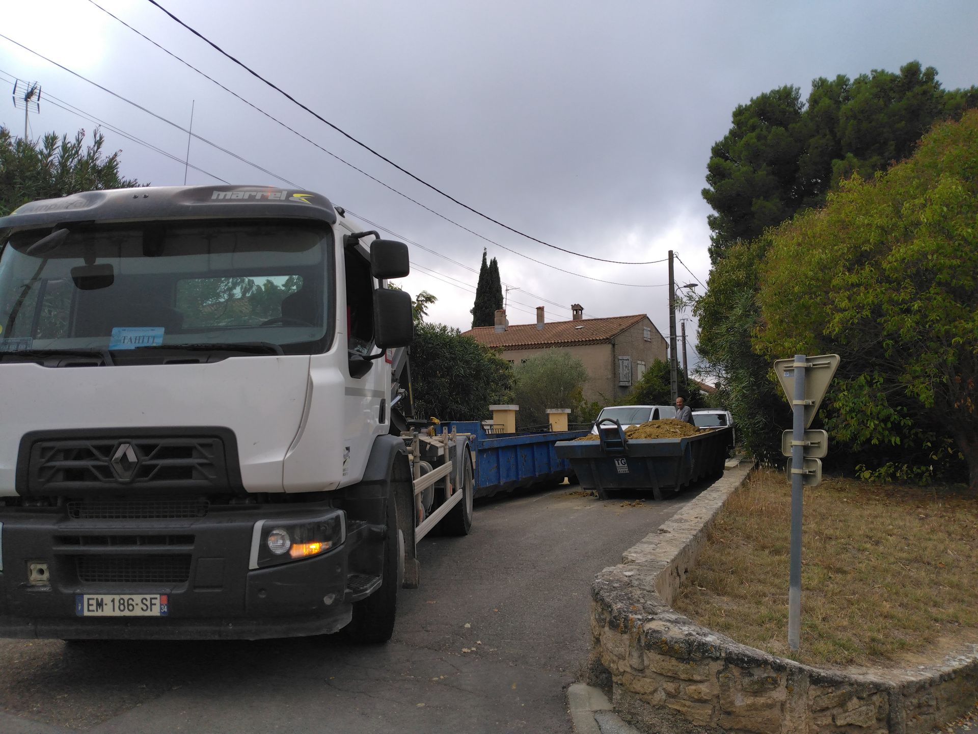 Un camion et sa remorque bloquent une rue résidentielle étroite près d'une maison et d'arbres.