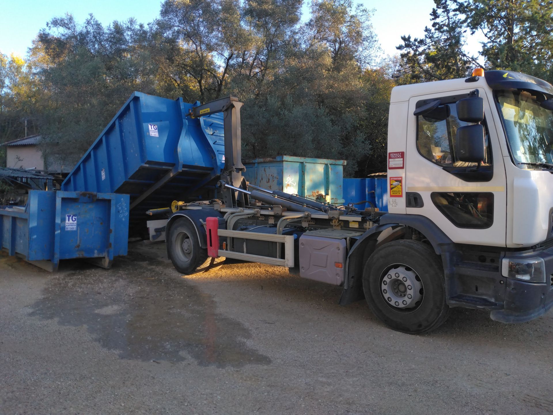 Un camion-benne bleu vide une benne à ordures dans sa trémie à l'extérieur, près des arbres et des bâtiments.