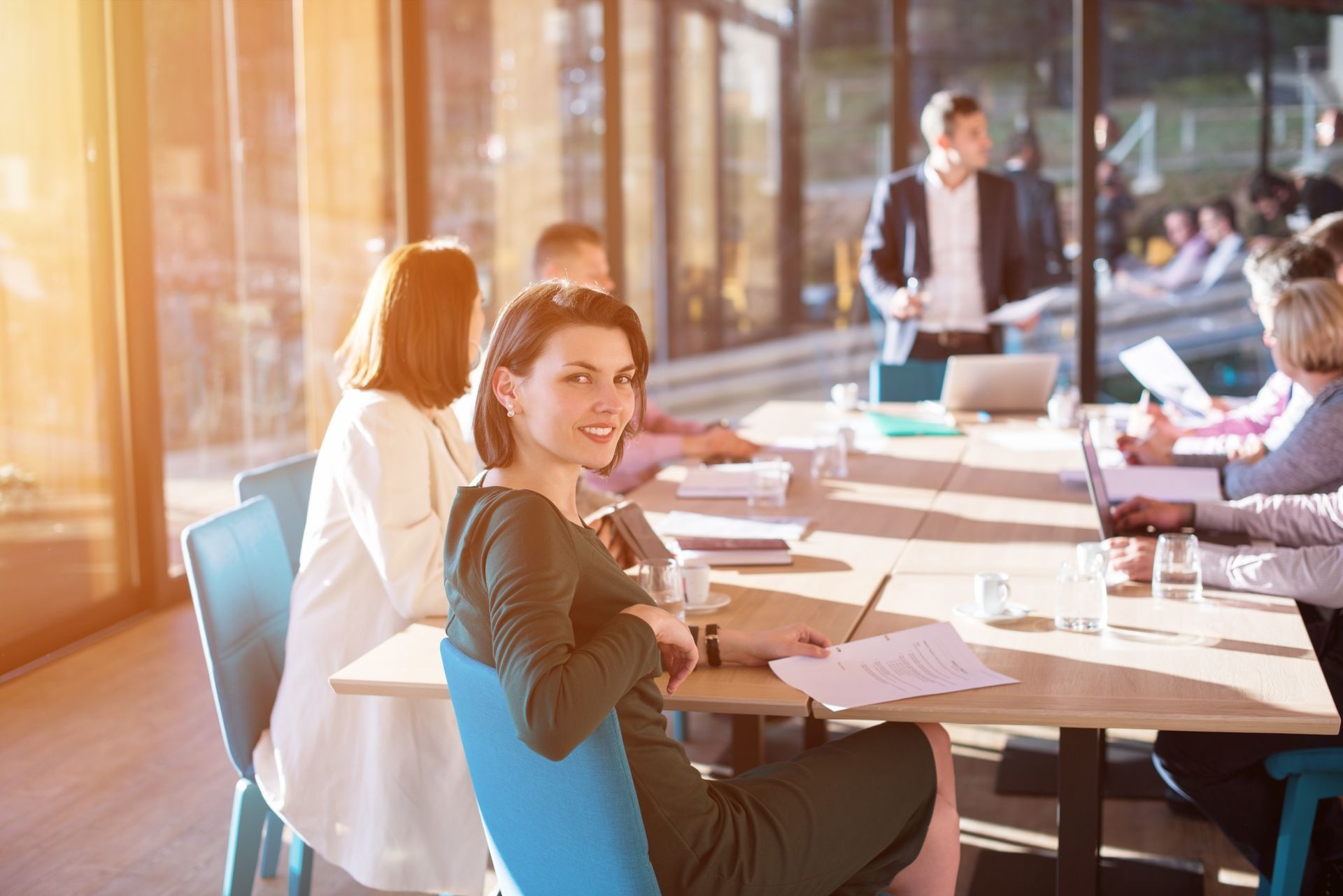 Reunión de negocios en curso; una mujer con vestido verde sonríe a la cámara. Otros están sentados alrededor de una mesa larga, y un hombre de pie, gesticulando.