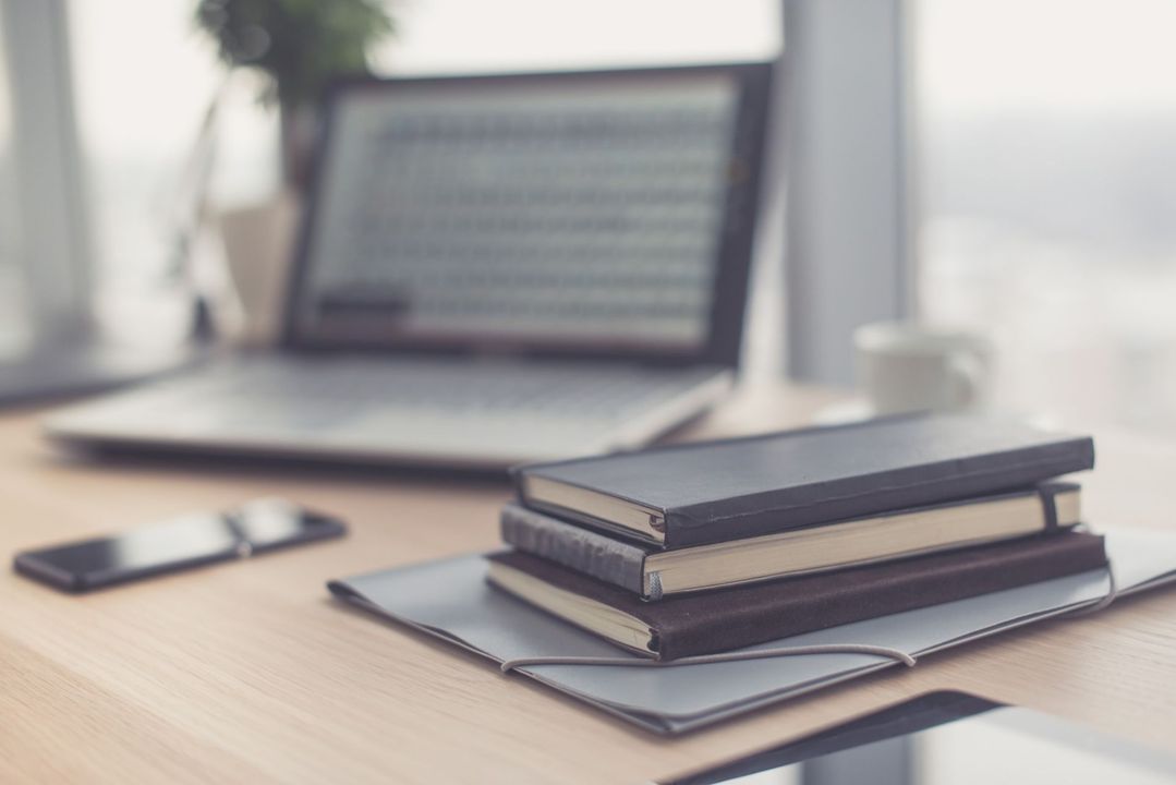 Computadora portátil, cuadernos y teléfono móvil sobre un escritorio junto a una ventana, lo que sugiere un espacio de trabajo.