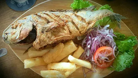 Un pescado frito con patatas fritas y verduras en un plato de madera.