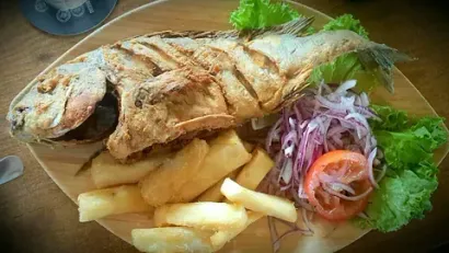 Un pescado frito con patatas fritas y verduras en un plato de madera.