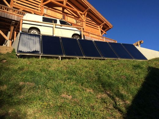 Des panneaux solaires alignés sur une pente herbeuse, à côté d'une camionnette blanche et d'un chalet en bois.