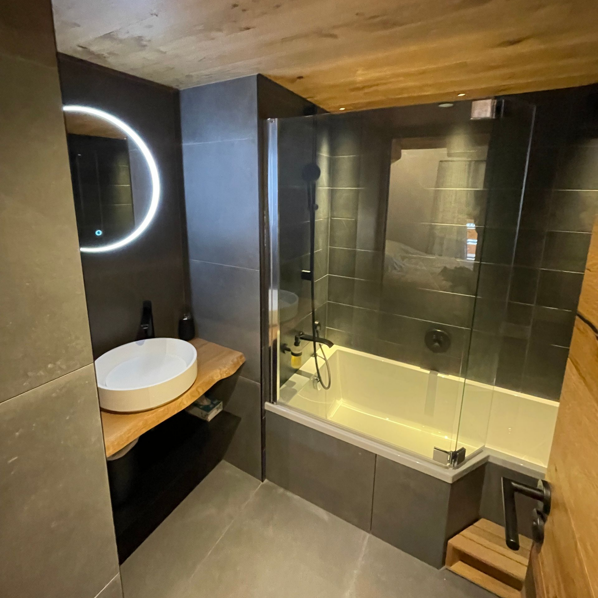 Salle de bains moderne noire et bois, avec douche en verre, vasque ronde, miroir rétroéclairé.