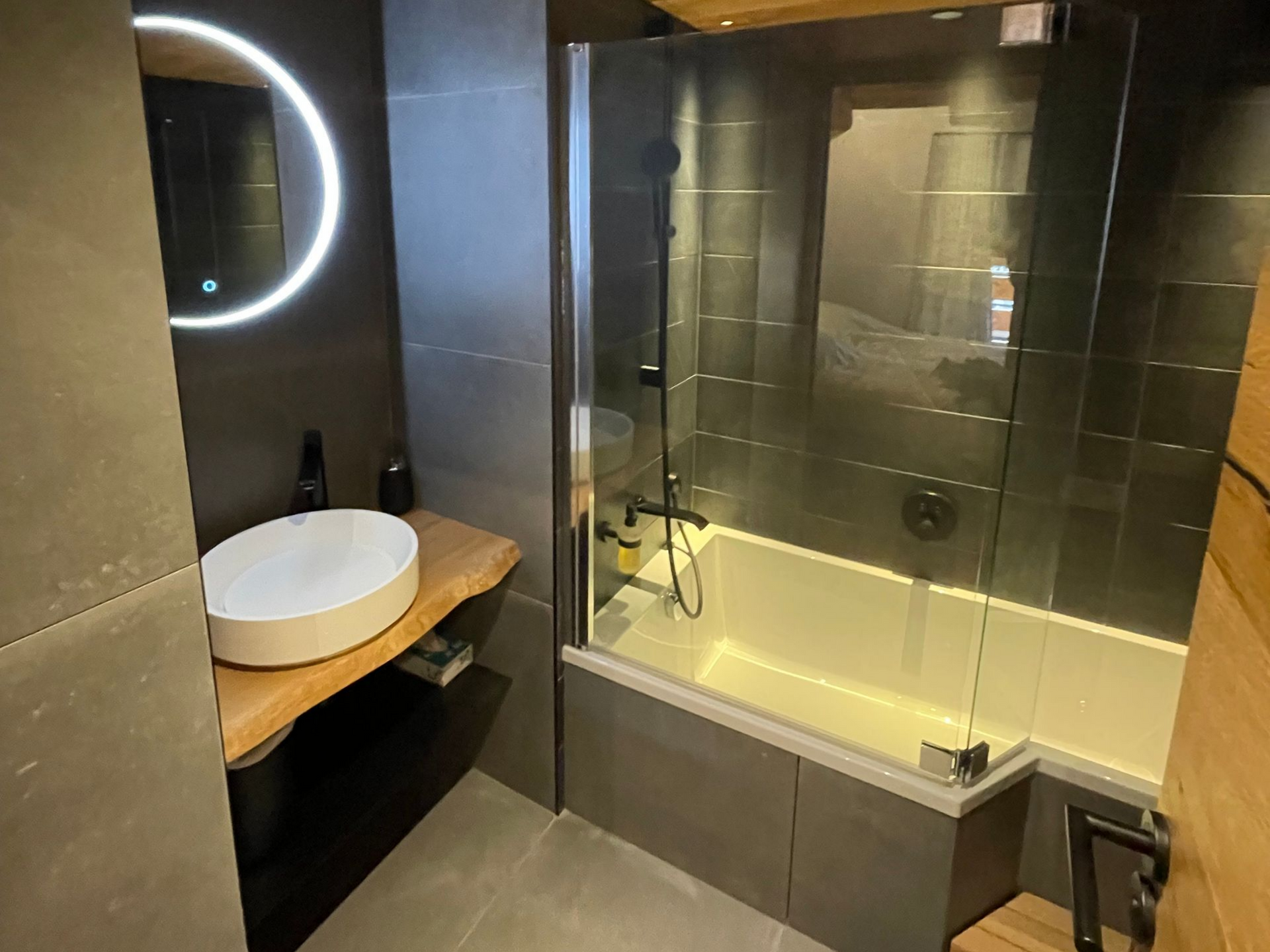 Salle de bains moderne avec miroir rond lumineux, lavabo et baignoire vitrée.