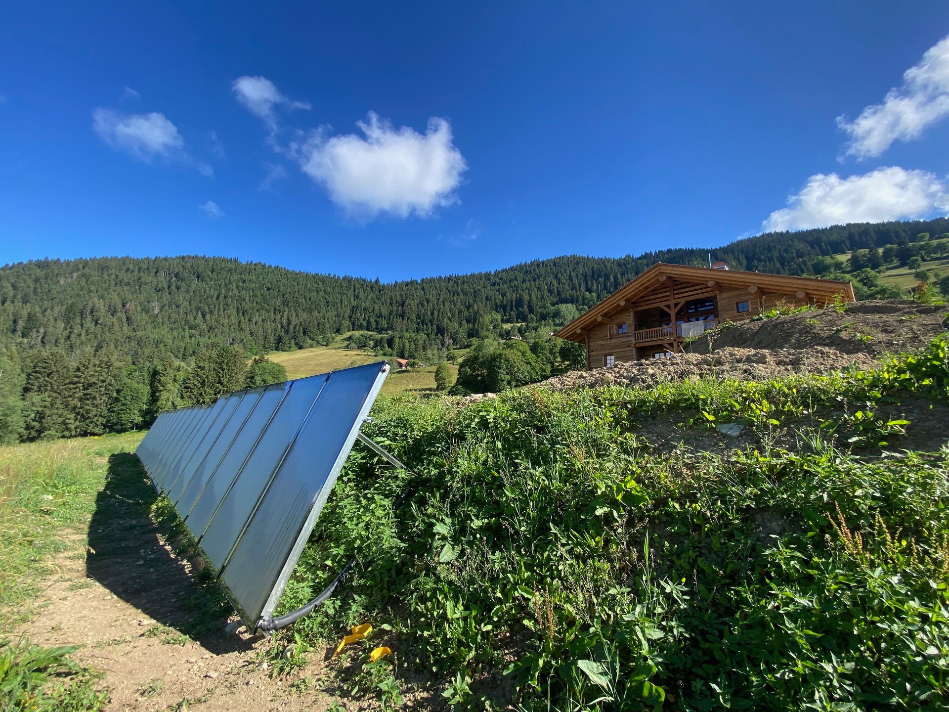 Des panneaux solaires dans un champ verdoyant à flanc de colline, avec une cabane et des montagnes boisées sous un ciel bleu.