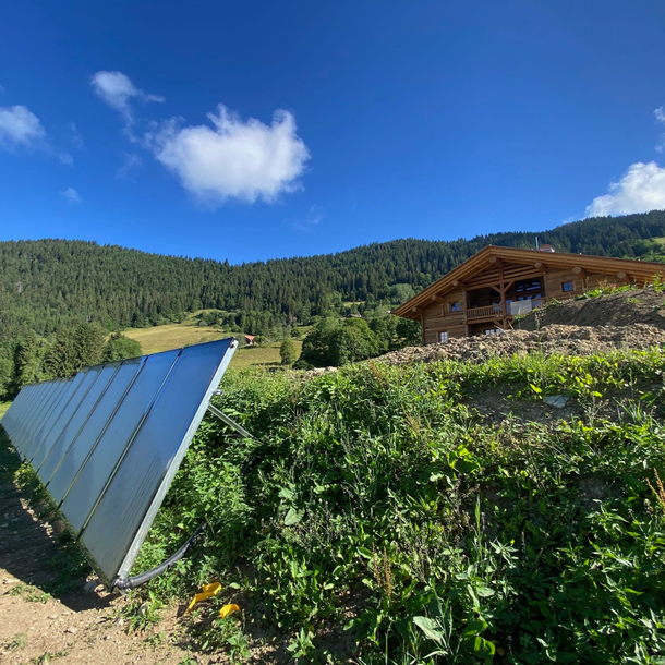Des panneaux solaires dans un champ verdoyant à flanc de colline, avec une cabane en bois sous un ciel bleu.