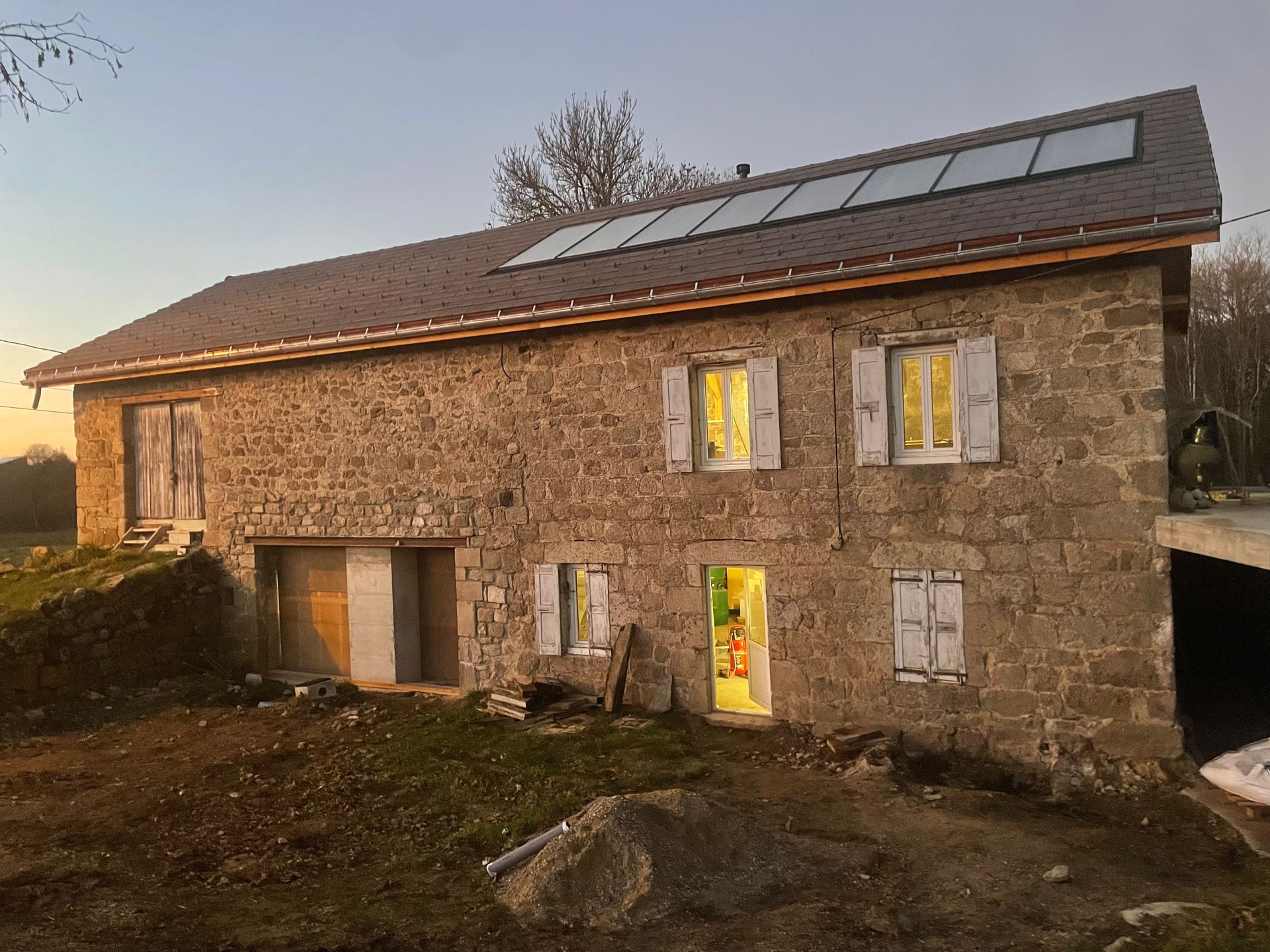 Maison en pierre au coucher du soleil, avec panneaux solaires sur le toit et fenêtres éclairées, vue depuis le jardin.