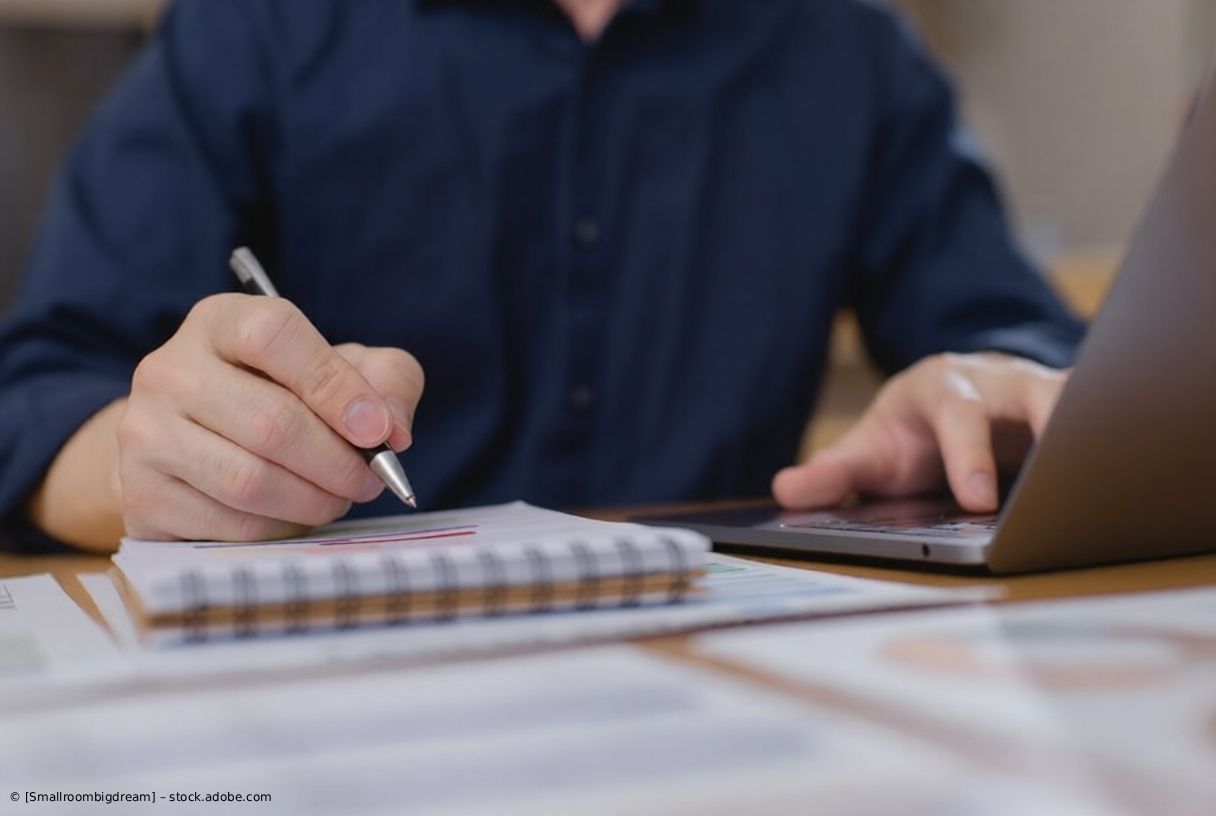 Eine Person schreibt in ein Notizbuch neben einem Laptop; Dokumente liegen auf dem Schreibtisch.