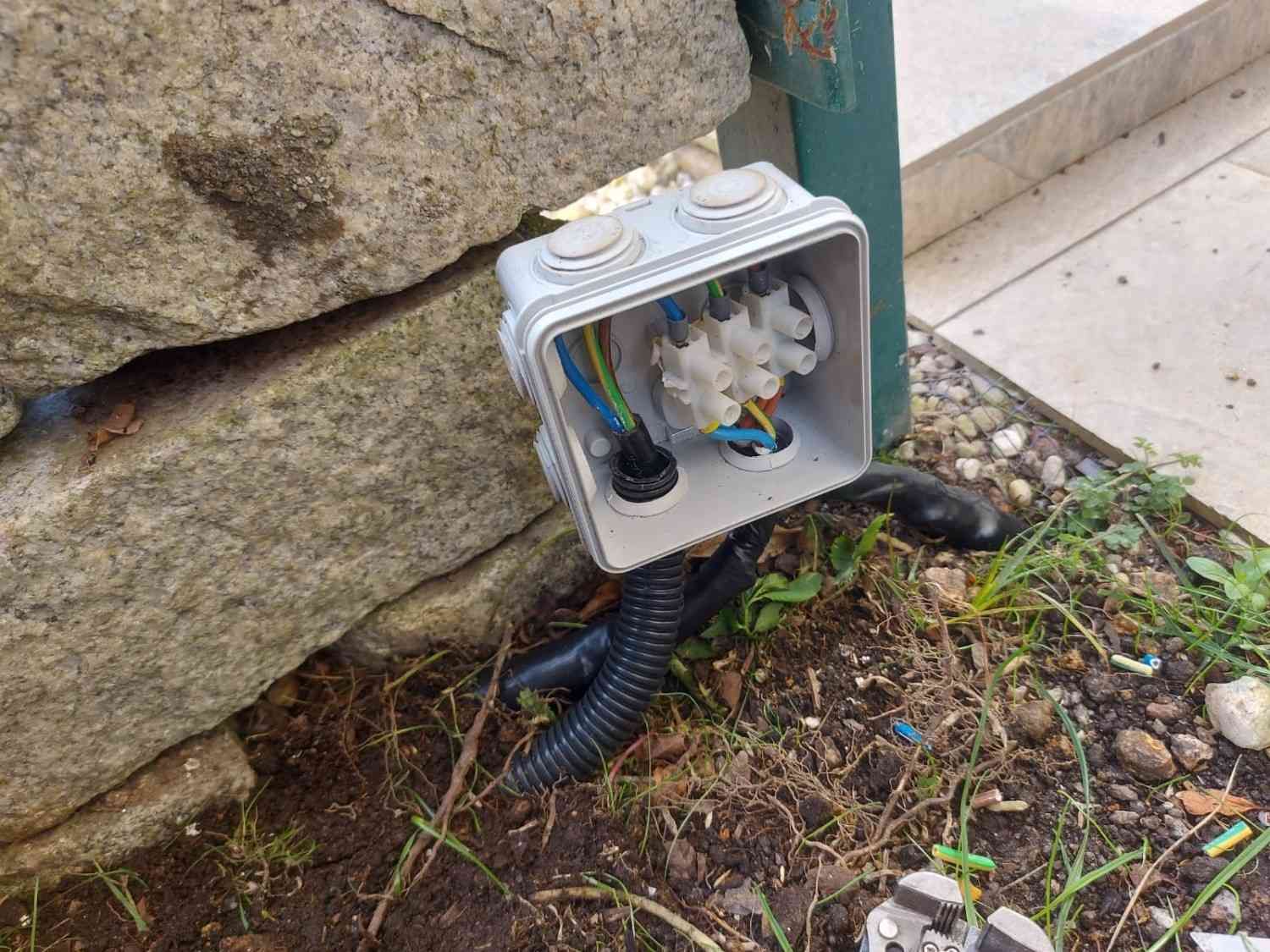 Caja eléctrica abierta con cables expuestos junto a un muro de piedra y la acera.