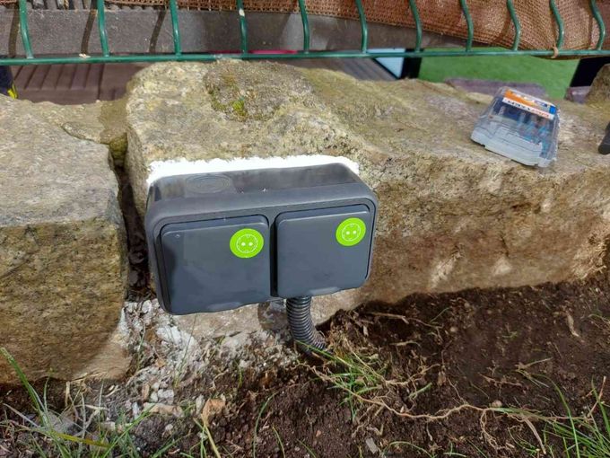 Dos cajas de servicios públicos grises montadas en un muro de piedra al aire libre, con pegatinas verdes en cada panel.