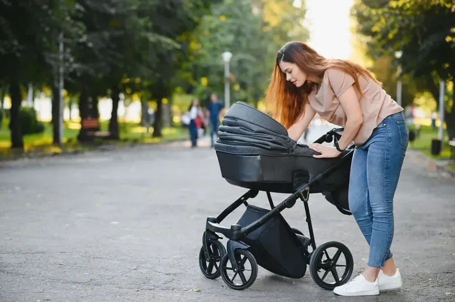 Mujer con jeans y una camiseta color canela mirando un cochecito negro en un camino pavimentado en un parque.