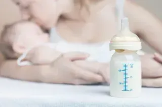 Biberón lleno de fórmula frente a una mujer que sostiene y besa a un bebé dormido.