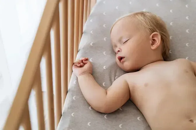 Bebé rubio durmiendo en una cuna de madera, acostado sobre una sábana estampada de color gris.