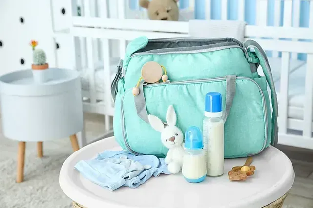 Una bolsa de pañales de color verde azulado con biberones, juguetes y ropa sobre una mesa en una guardería.
