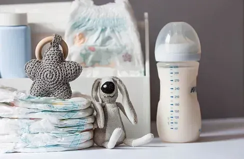 Elementos esenciales para bebés: pañales apilados, biberón de fórmula, juguetes de peluche y un paquete de pañales.