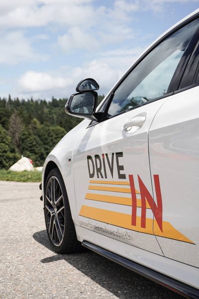 Voiture blanche d'auto-école avec le logo « DRIVE IN », garée à l'extérieur sur une route près des arbres et des montagnes