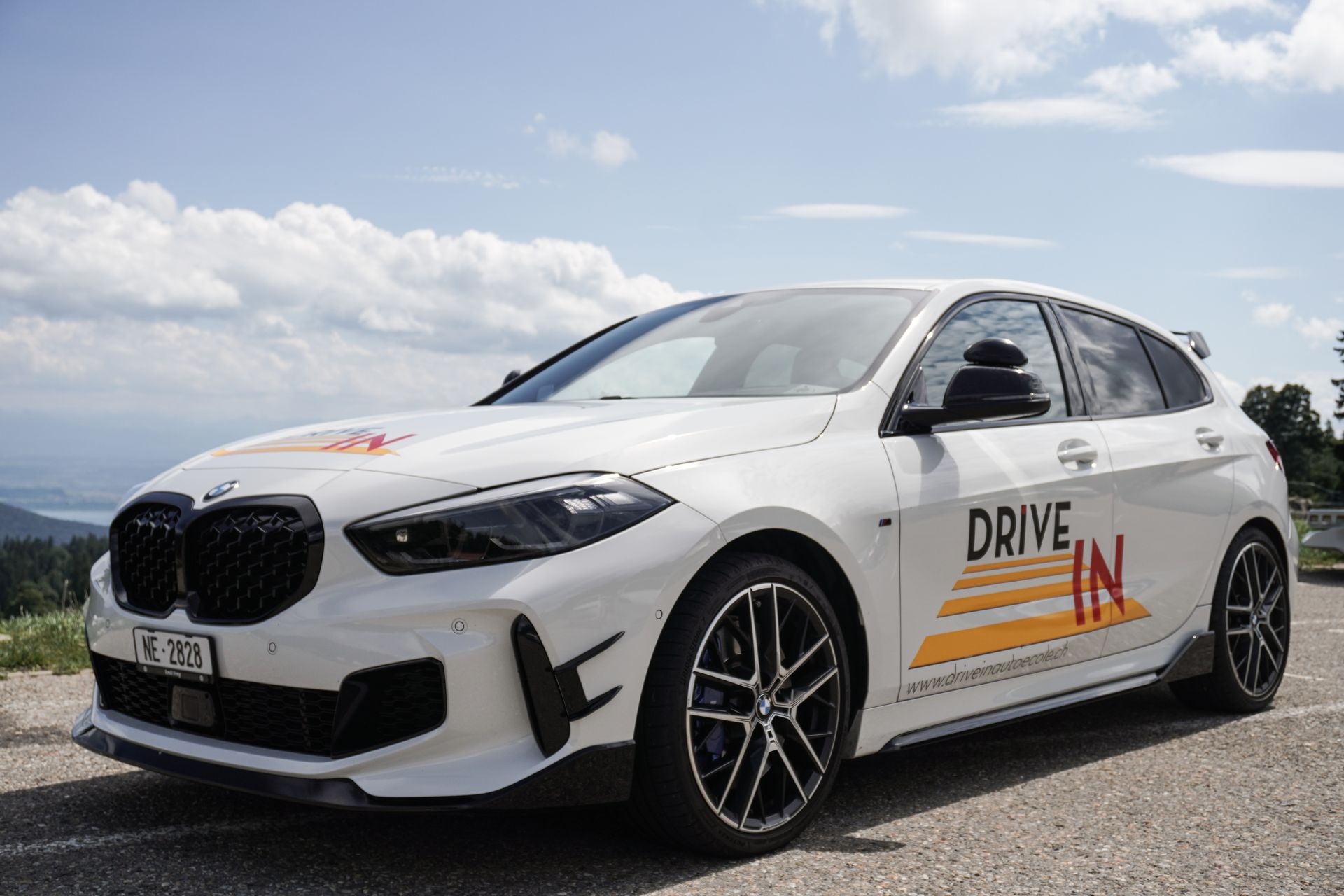 Une BMW blanche à hayon avec le logo « DRIVE » est garée à l'extérieur sous un ciel nuageux.