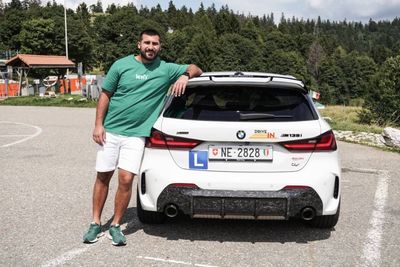 Un homme appuyé contre un SUV BMW blanc sur une route de montagne, avec des arbres et des bâtiments en arrière-plan.