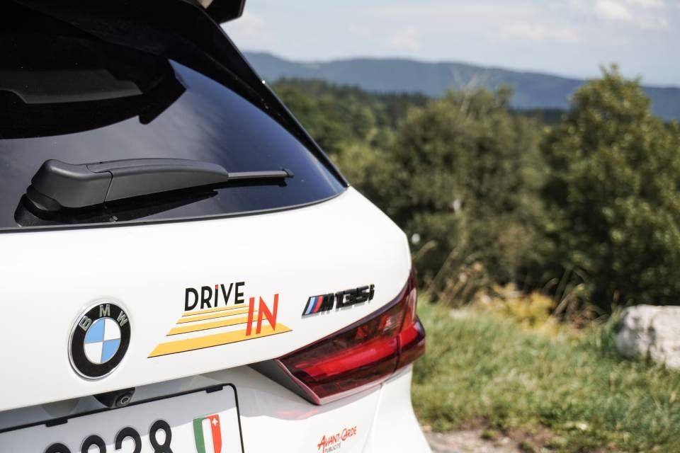 Arrière d'une BMW M8 blanche avec un autocollant « Drive IN », garée au bord d'une route de montagne pittoresque.