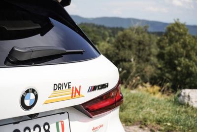 Arrière d'une BMW M8 blanche avec un autocollant « Drive IN », garée au bord d'une route de montagne pittoresque.
