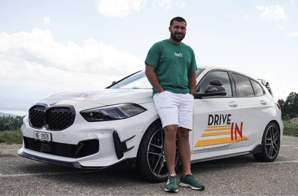 Un homme se tient debout à côté d'une voiture blanche à hayon avec l'inscription « DRIVE IN » sur la portière, sur un parking gravillonné sous un ciel nuageux.