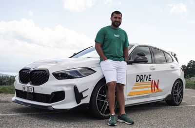 Un homme se tient debout à côté d'une voiture blanche à hayon avec l'inscription « DRIVE IN » sur la portière, sur un parking gravillonné sous un ciel nuageux.