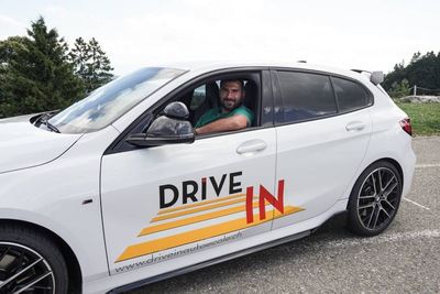 Voiture blanche avec logo « DRIVE IN », conducteur souriant au volant sur un parking