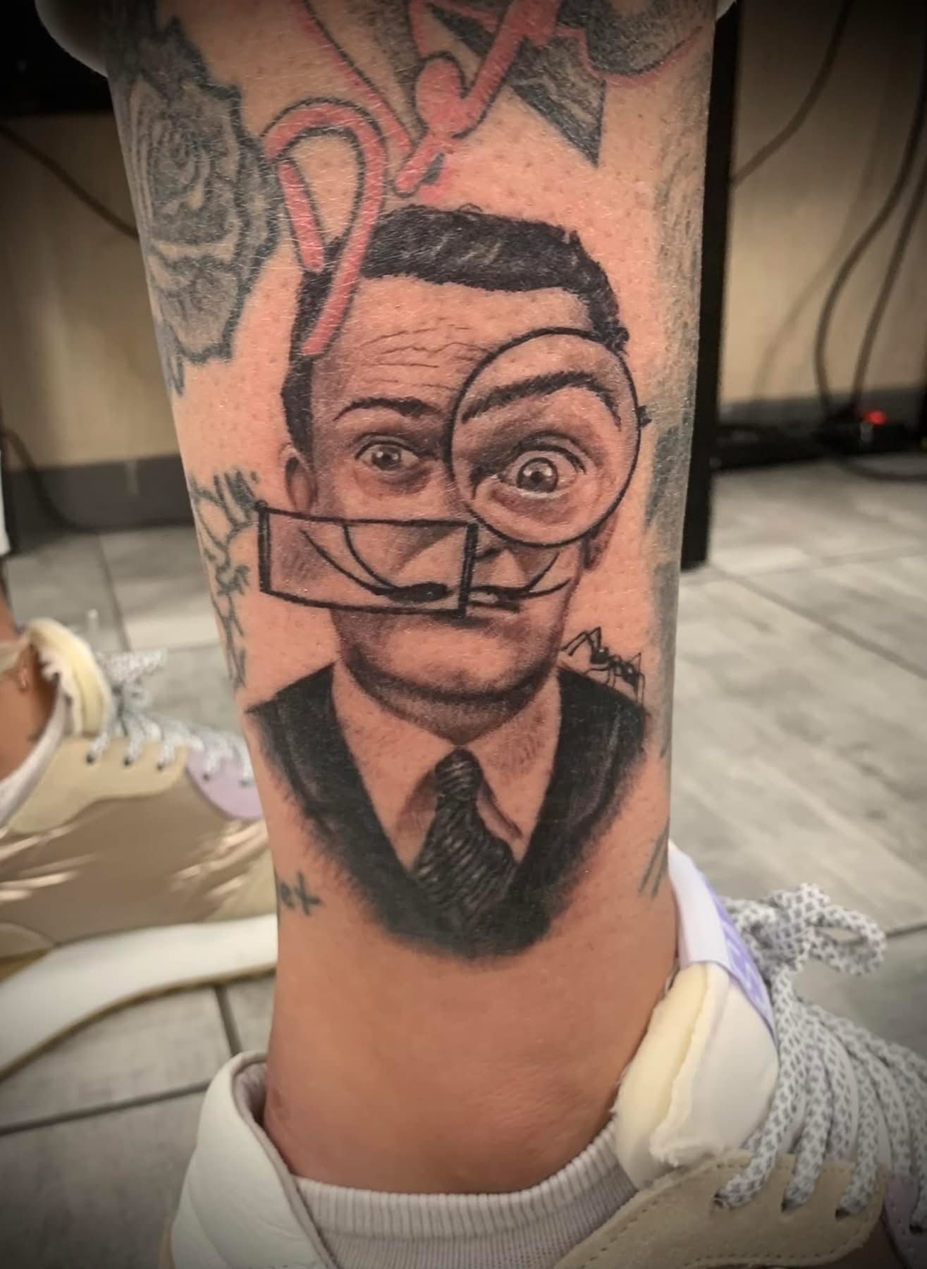Un tatouage sur la jambe représentant un homme regardant à travers une loupe, avec un élément rectangulaire surréaliste au-dessus de sa bouche.