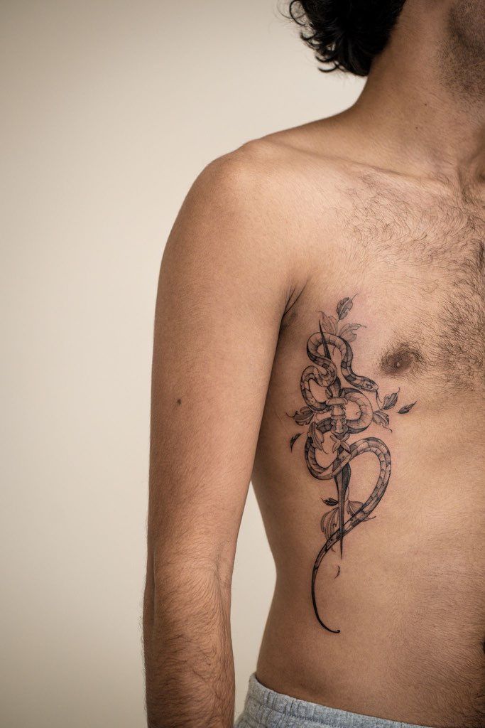 Le torse d'une personne présentant un tatouage à l'encre noire représentant un serpent enroulé autour d'un poignard, avec des accents de roses sur la cage thoracique.
