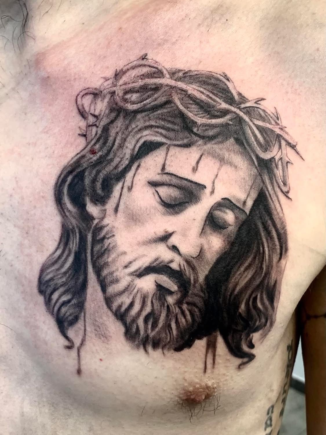 Un tatouage en noir et gris sur la poitrine d'une personne, représentant le visage triste de Jésus portant une couronne d'épines.