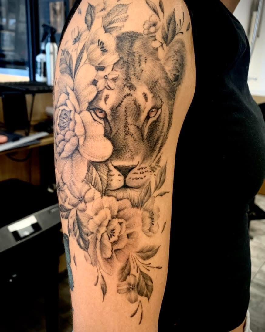 Un tatouage sur le haut du bras représentant un visage de lion réaliste entouré de motifs floraux détaillés à l'encre noire et grise.