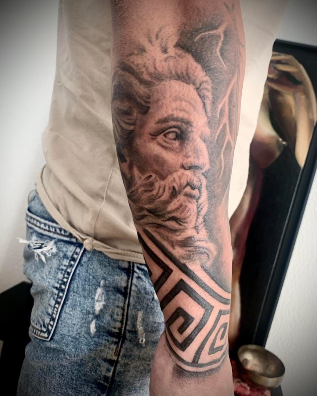 Un tatouage en noir et gris représentant une divinité classique sur le haut du bras, avec un portrait et une bordure à motif grec.