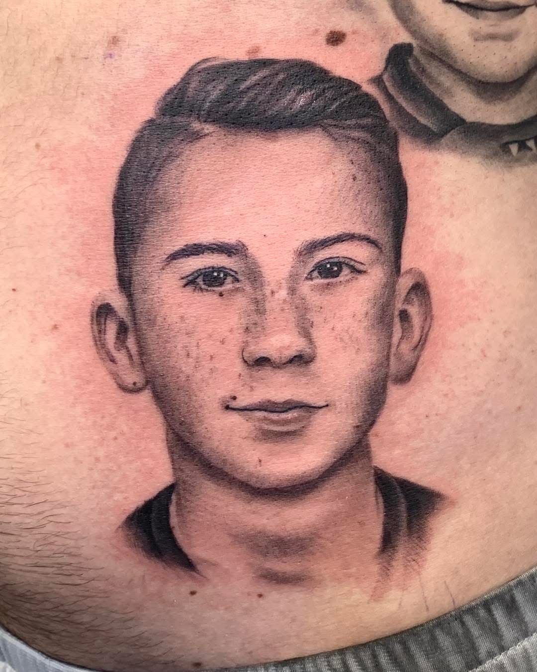 Un tatouage réaliste représentant le portrait d'une jeune personne sur la peau, avec des ombrages détaillés, une coiffure texturée et des taches de rousseur.
