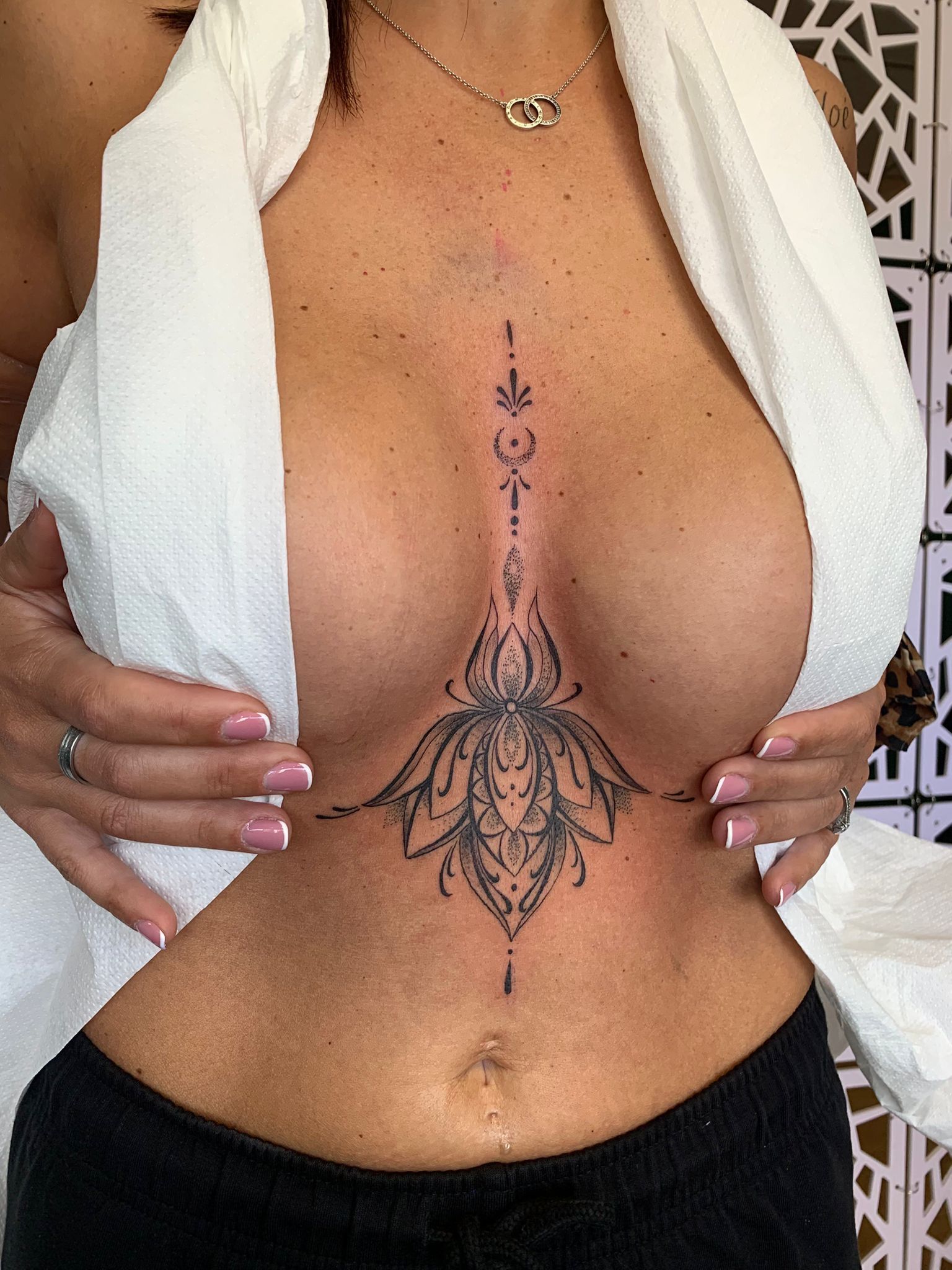 Gros plan sur un tatouage du sternum présentant des motifs complexes de mandala et de fleurs à l'encre noire, centrés entre les seins.