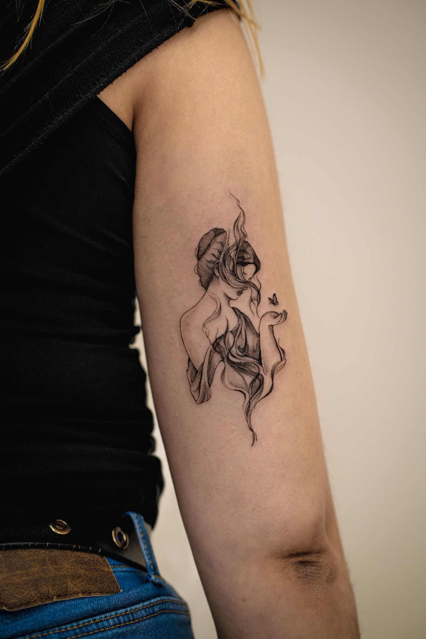 Gros plan sur le haut du bras d'une personne présentant un délicat tatouage à l'encre noire représentant une figure abstraite et éthérée.