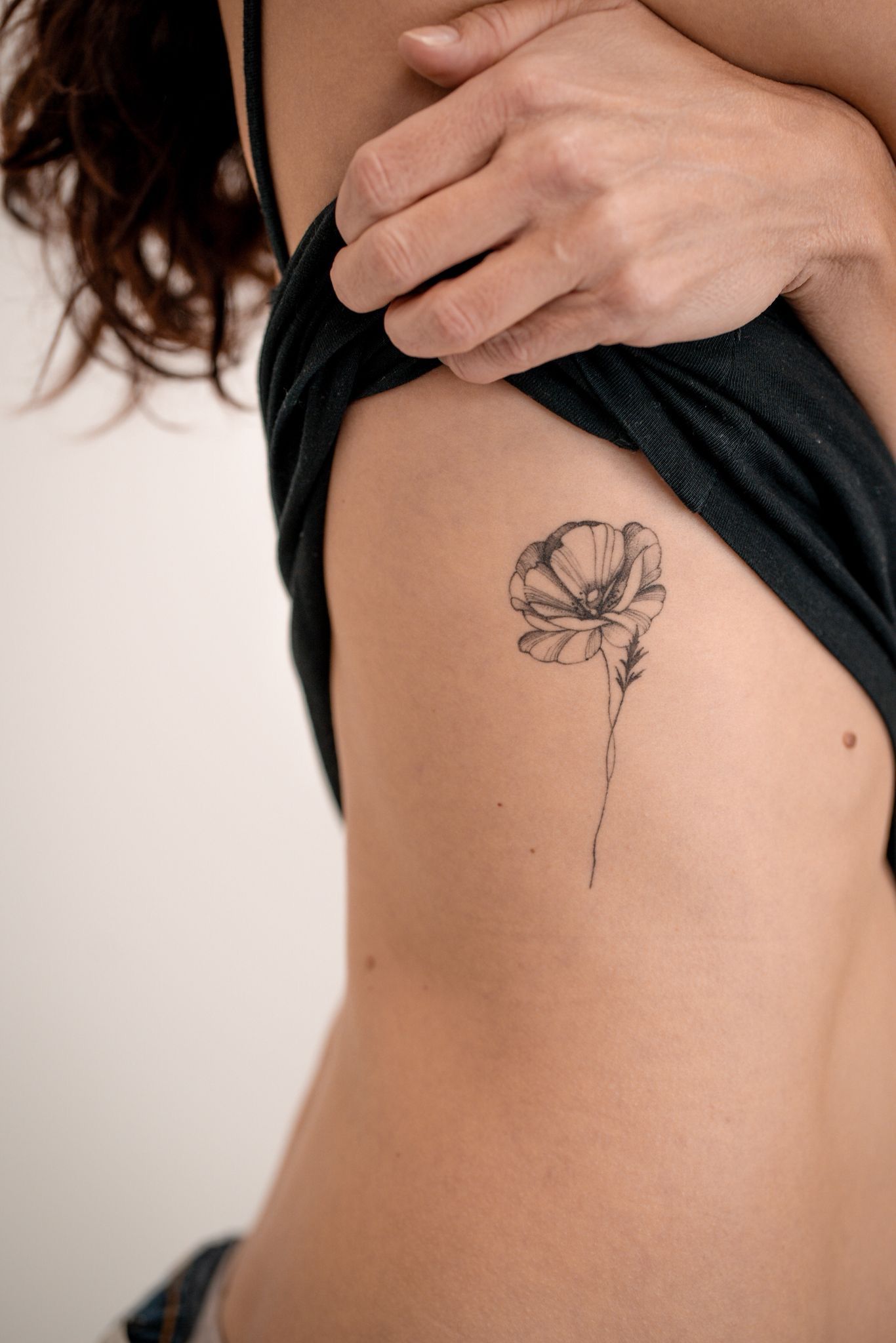Un tatouage floral délicat aux lignes fines, ornant le flanc du torse, représentant une fleur esquissée à la tige minimaliste.