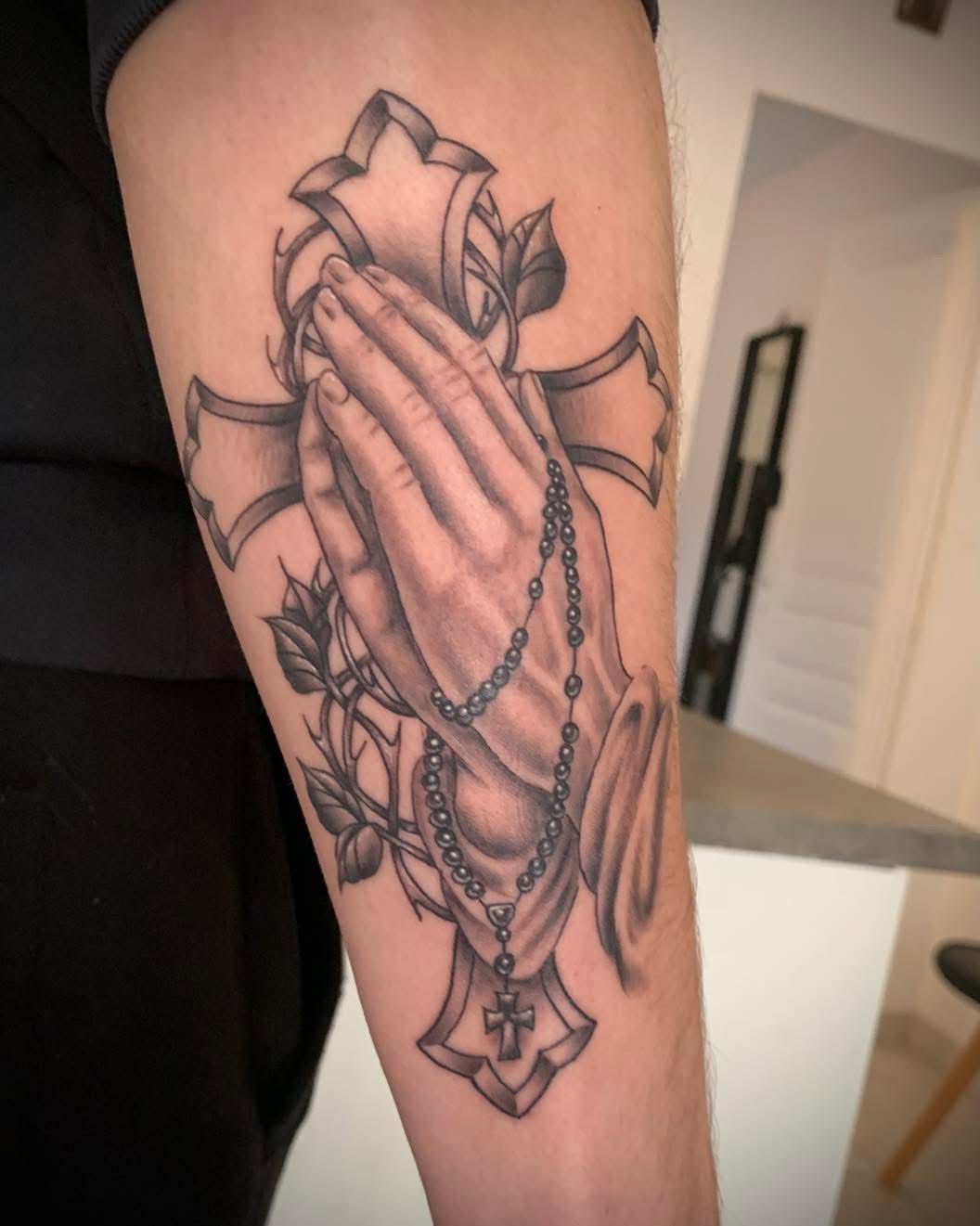 Un tatouage en noir et gris sur un bras représentant des mains jointes en prière tenant un chapelet devant une croix ornée de motifs floraux.