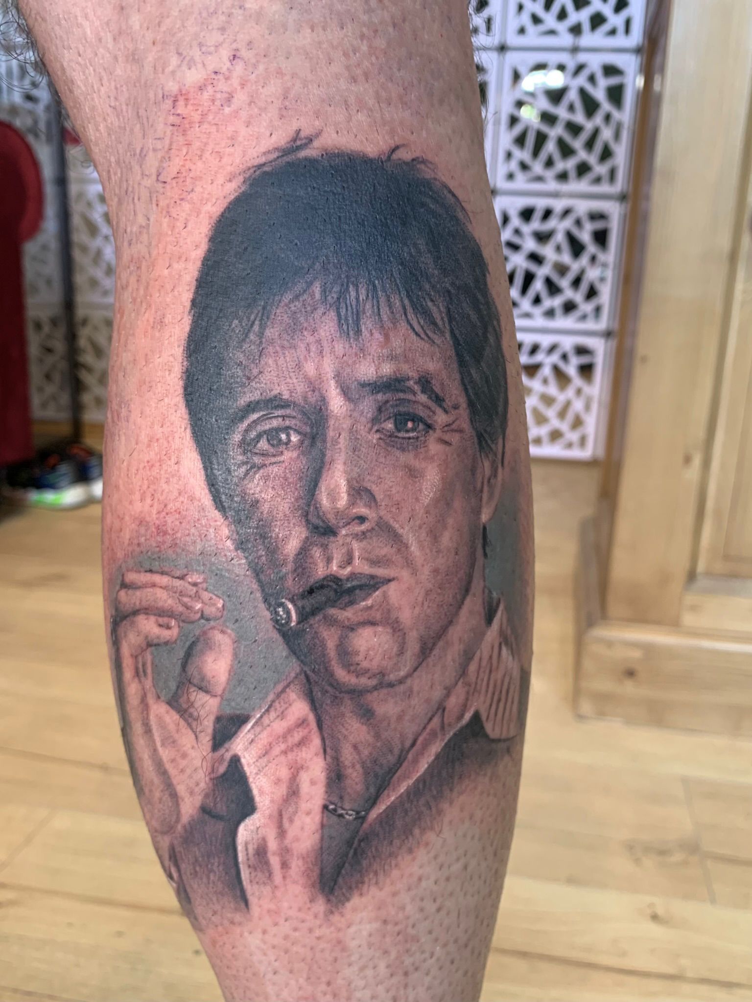 Un tatouage en noir et gris sur la jambe d'une personne représentant Al Pacino dans le rôle de Tony Montana du film Scarface, tenant un cigare.