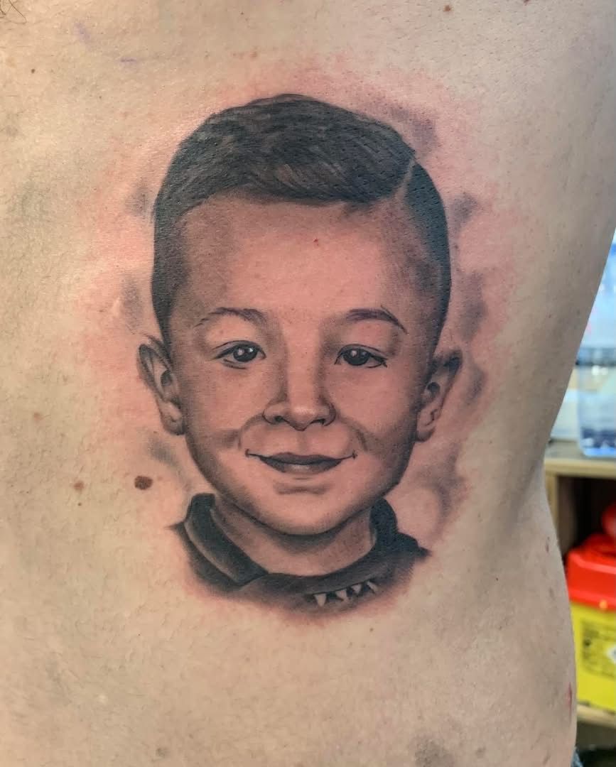 Tatouage portrait en noir et gris sur la peau d'un enfant aux cheveux courts et au doux sourire, entouré d'ombres claires.