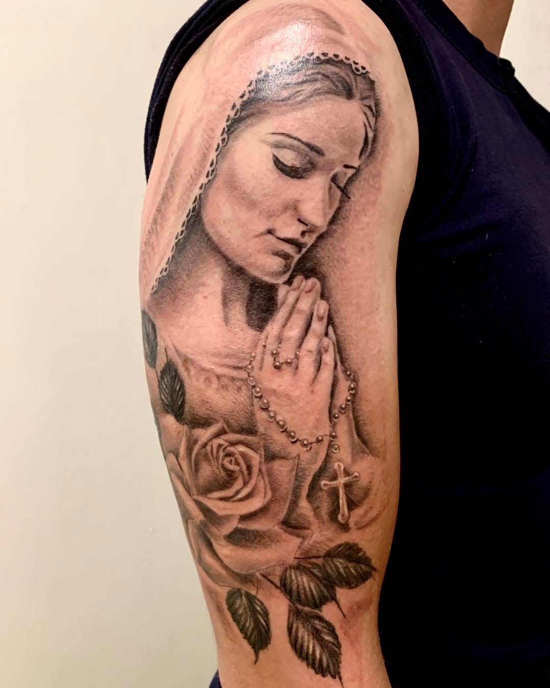 Tatouage noir et gris sur le haut du bras représentant une femme aux yeux fermés, les mains jointes en prière, un chapelet et des roses.