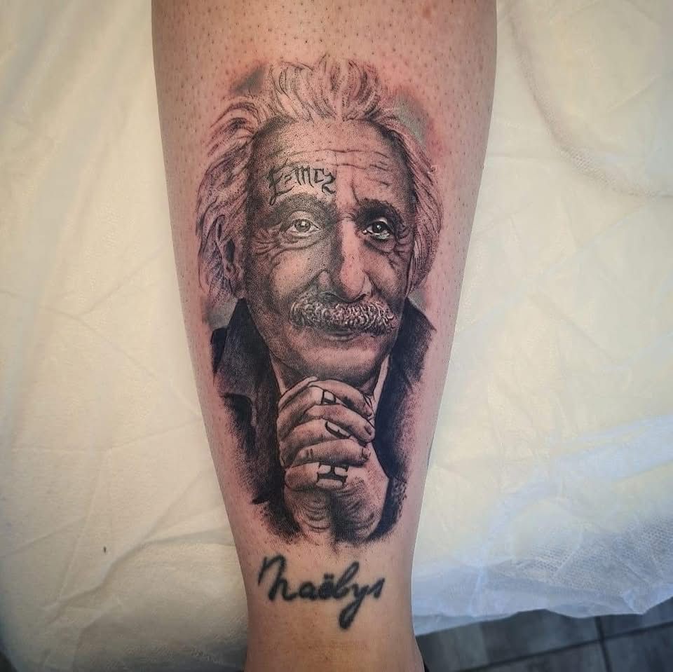 Tatouage en noir et blanc représentant Albert Einstein, les mains jointes, une inscription sur le front et un nom en dessous.