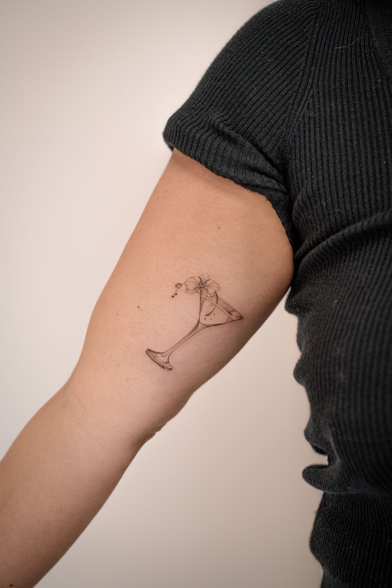 Un petit tatouage aux lignes fines représentant un verre à martini avec une garniture, situé sur la partie supérieure interne du bras d'une personne.