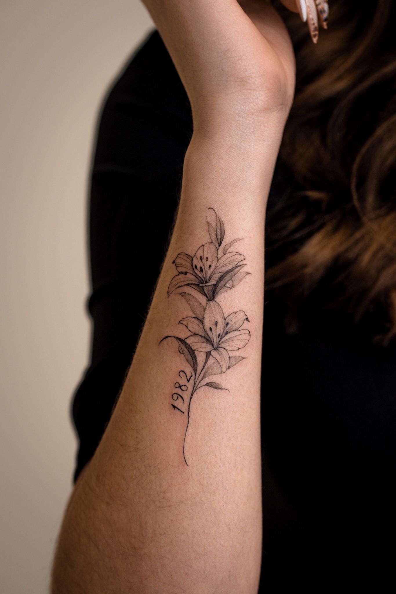Gros plan sur un avant-bras orné d'un tatouage fin à l'encre noire représentant deux fleurs de lys avec le nombre « 1982 » inscrit.