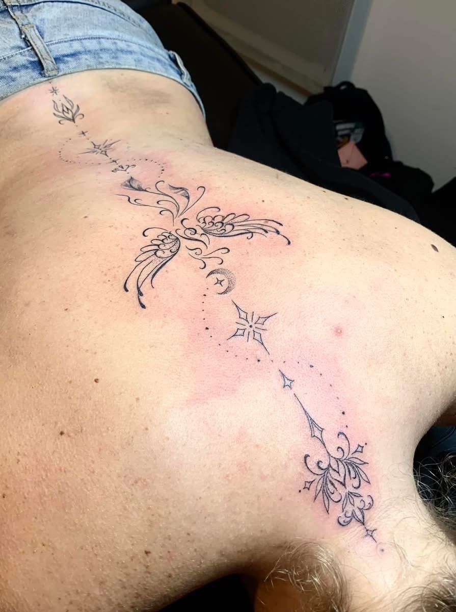 Un tatouage fin le long de la colonne vertébrale, orné de filigranes décoratifs, de symboles d'étoiles et de points délicats.