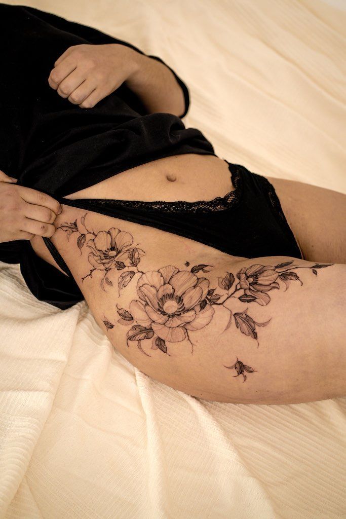 Un tatouage floral sur le haut de la cuisse, représentant des fleurs noires et blanches détaillées et une petite abeille sur fond blanc.