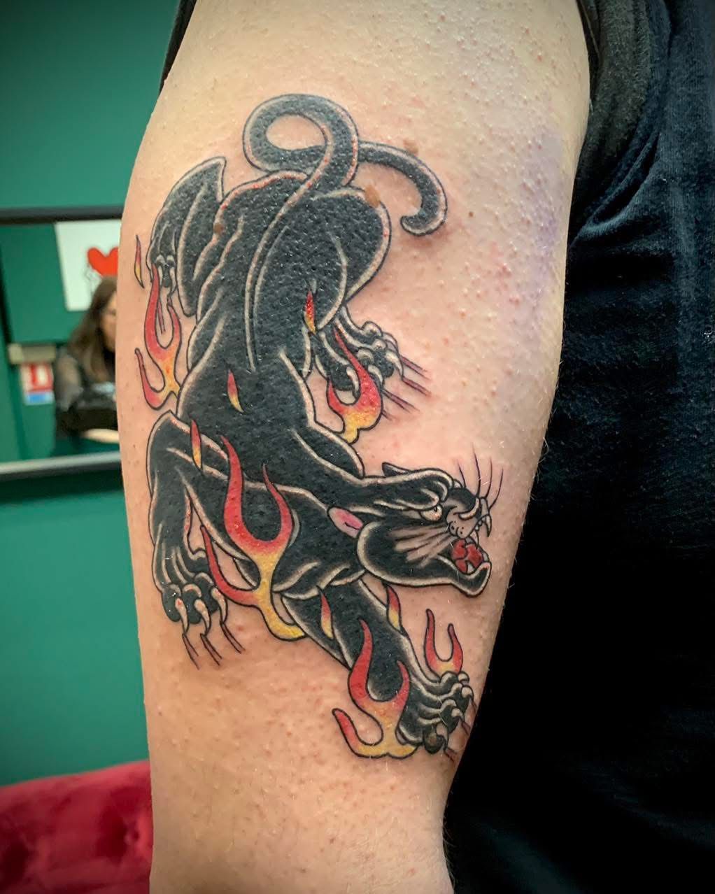 Un tatouage de style traditionnel sur le haut du bras représentant une panthère noire bondissant à travers des flammes stylisées orange et jaunes.
