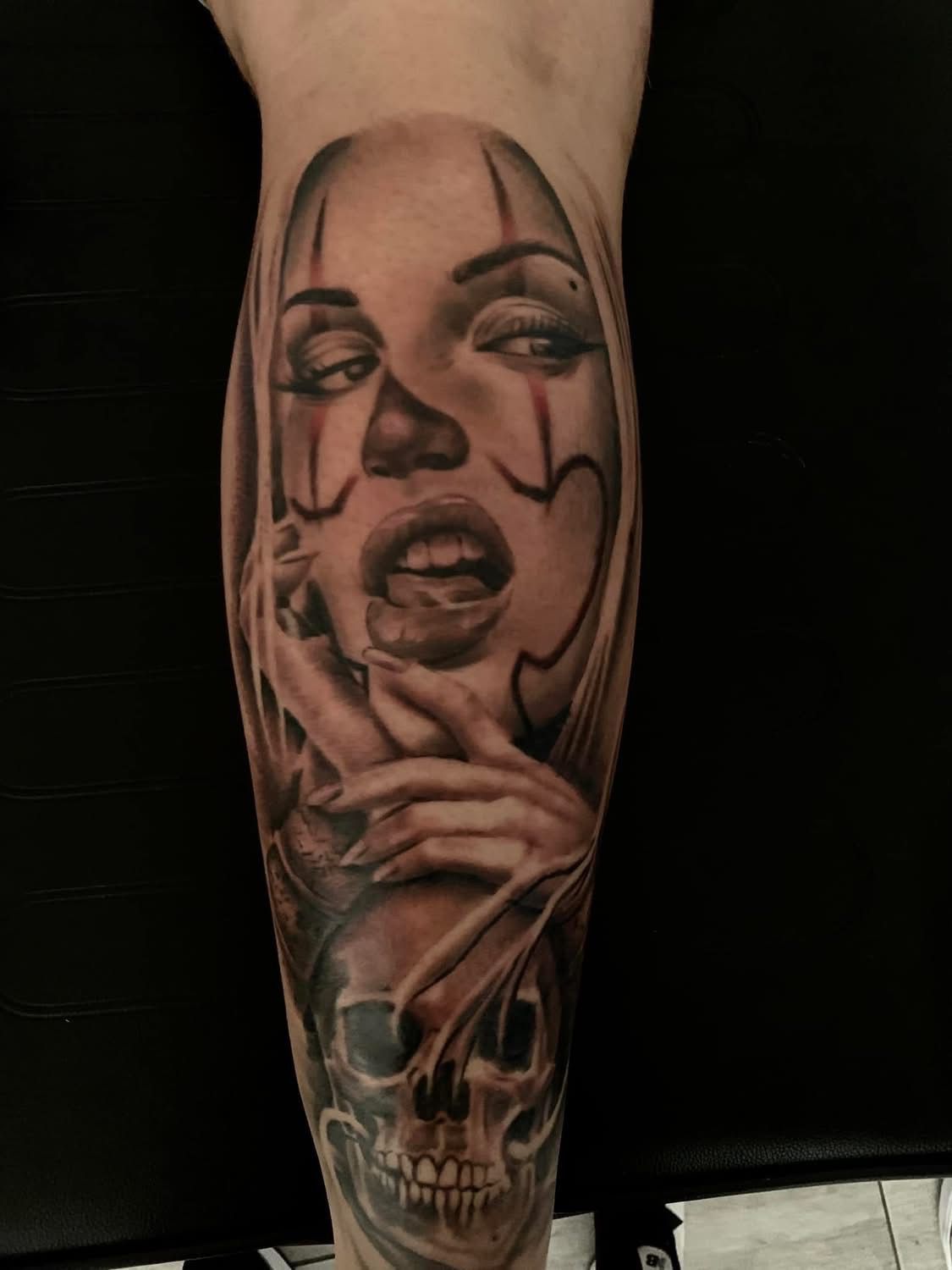 Tatouage noir et gris sur un membre représentant un visage de femme stylisé avec un maquillage de clown au-dessus d'un crâne humain détaillé.