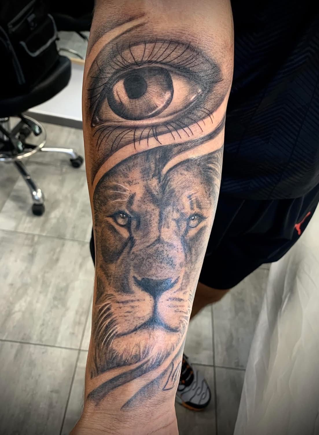 Un tatouage en noir et gris sur l'avant-bras d'une personne, représentant un grand œil réaliste au-dessus du visage d'un lion.