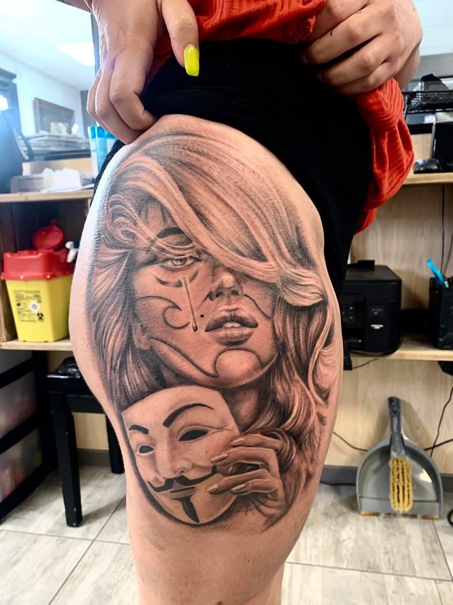 Un tatouage en noir et gris sur la cuisse d'une personne, représentant le visage d'une femme partiellement dissimulé par un masque de Guy Fawkes.
